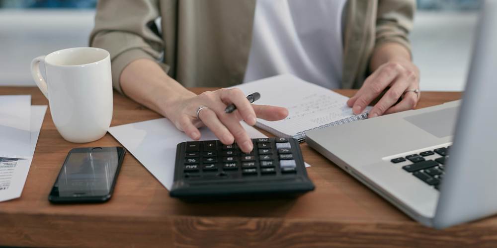 Przeliczanie kosztów na kalkulatorze i laptopie. Obok na biurku smartfon oraz biały kubek z kawą.