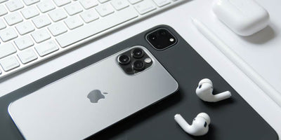 Na zdjęciu iPhone, a pod nim iPad w czarnym kolorze oraz białe słuchawki AirPods. W prawym górnym rogi etui do słuchawke w kolorze białym oraz biała klawiatura.