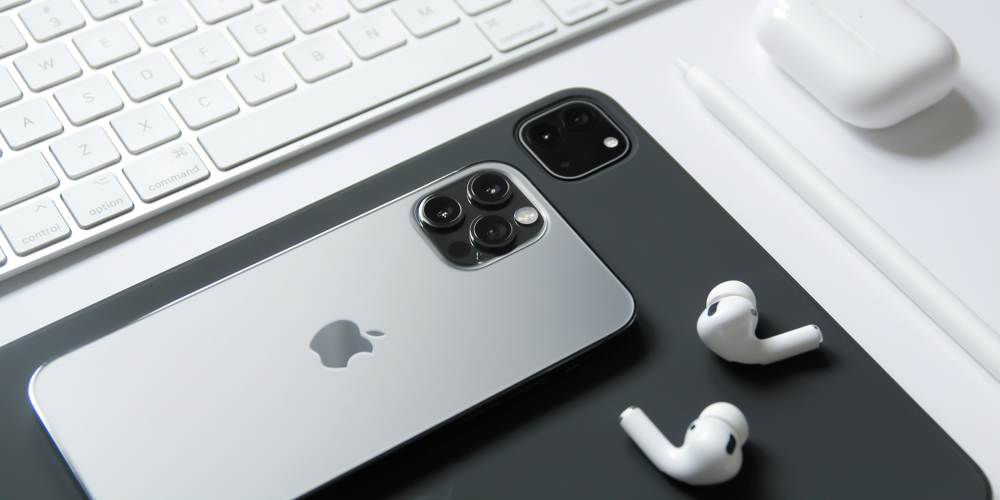 Na zdjęciu iPhone, a pod nim iPad w czarnym kolorze oraz białe słuchawki AirPods. W prawym górnym rogi etui do słuchawke w kolorze białym oraz biała klawiatura.